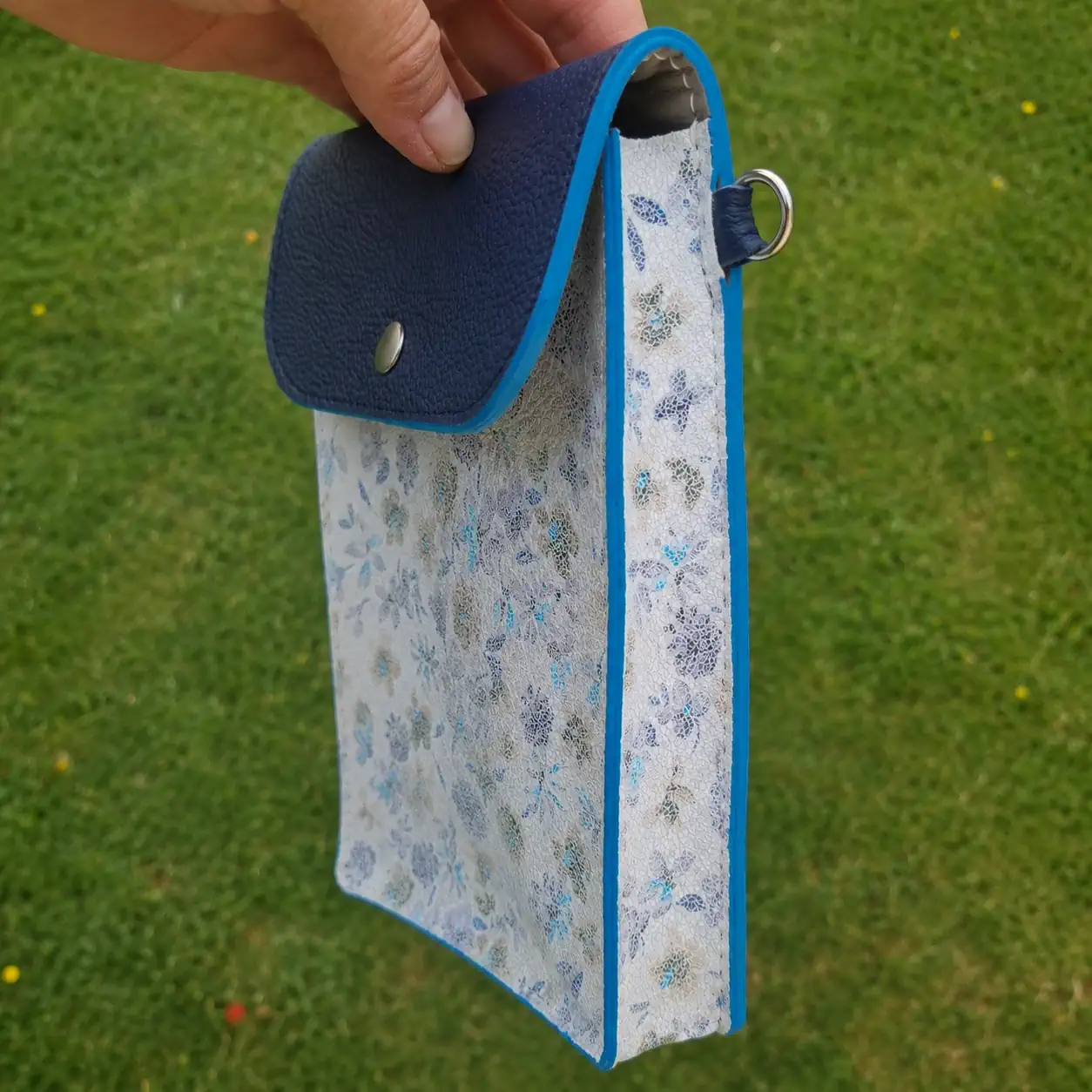 Pochette en cuir pratique chic printanière, de couleur bleu marine et blanche avec des motifs à fleurs bleues. Pour téléphone, lunettes et portefeuille. Portée en bandoulière. Profil.